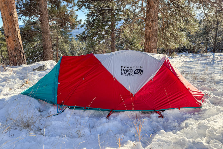 The Mountain Hardwear Trango 2 tent fully pitched in snow, showing its red and teal fly with the logo centered on the white panel.