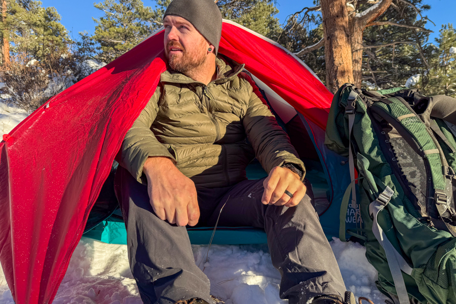 A person sitting inside the Trango 2’s open vestibule on a snowy morning, wearing a puffy jacket and looking off to the side with a backpack beside them.