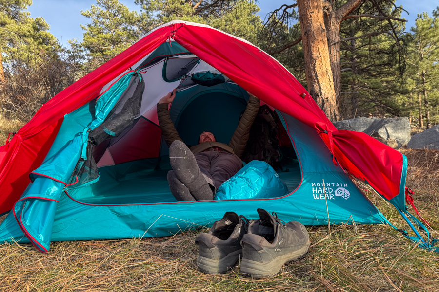 A person lying on their back inside the Trango 2 tent, reaching up toward the ceiling, with the red fly pulled back and shoes resting just outside the entrance.