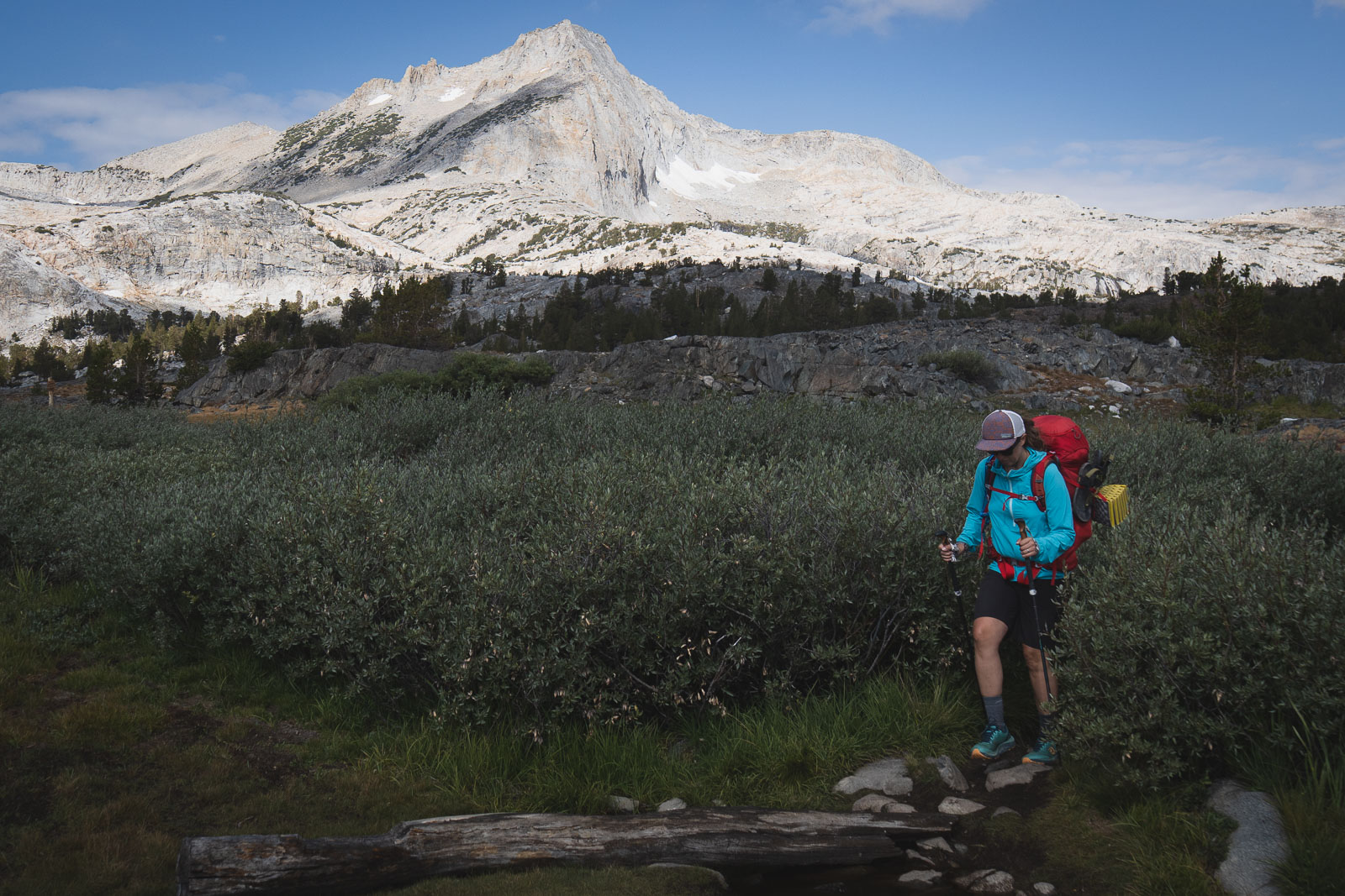Pregnant person backpacking, crossing a stream, with a granite mountain in the background.