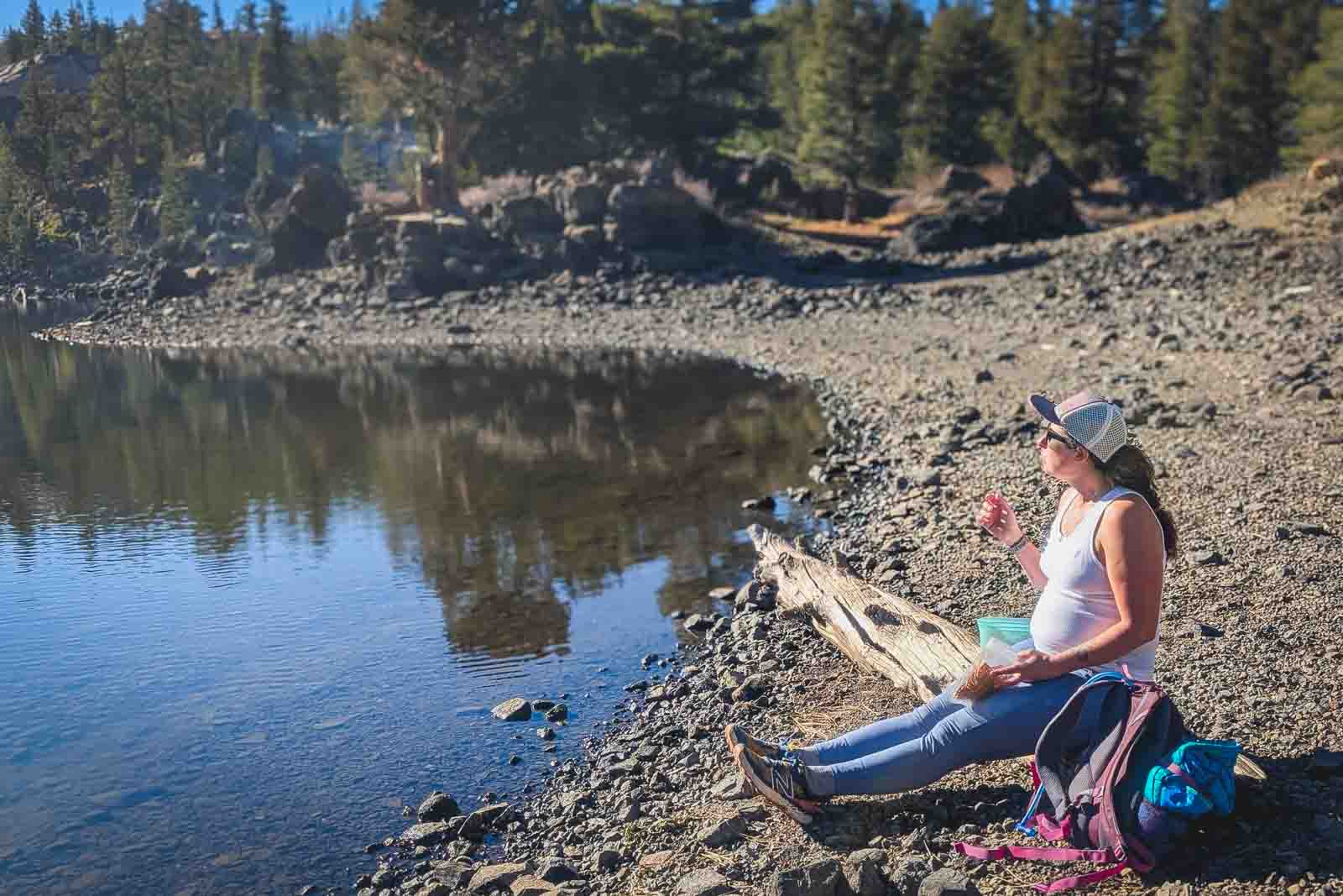 Pregnant person sitting on a log next to a lake, looking at the view, while eating snacks.