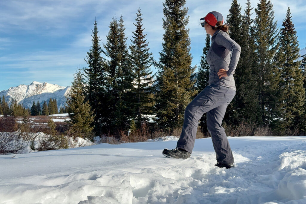 A person wearing the Toundra boots stands among some trees and snow admiring a view of the mountains.