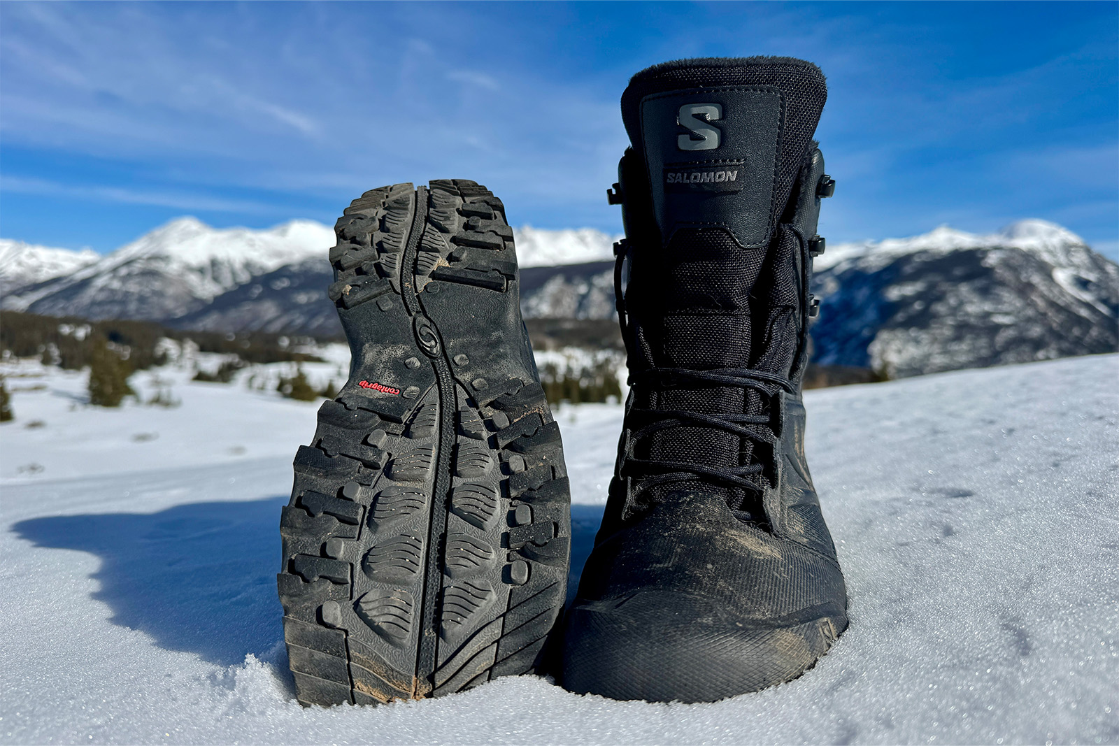 A Toundra boot sits in the snow. Next to it another boot is turned over to show the tread. There are mountains in the background.