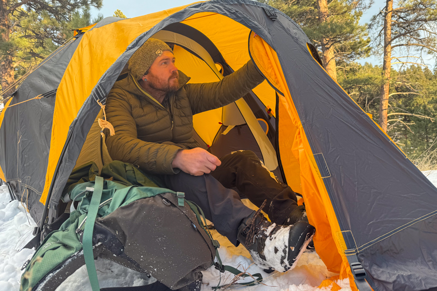 A person sitting inside the open vestibule of the Mountain 25 tent, adjusting the door while snow-covered boots and a packed backpack sit outside on the snow.