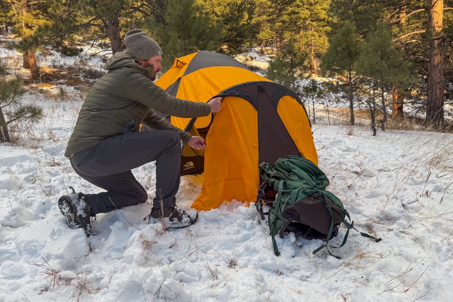 A person kneeling in the snow while zipping the vestibule door of the Mountain 25 tent closed, with a backpack resting beside the entrance.