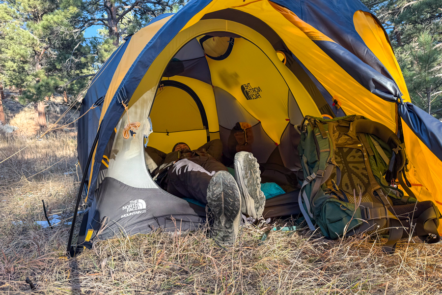 A person lying on their back inside The North Face Mountain 25 tent with the vestibule open, boots resting in the doorway and a backpack beside the entrance.
