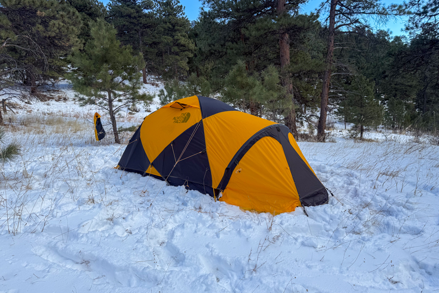 Side view of the Mountain 25 tent in snow, showing the yellow-and-black fly, a domed vestibule, and surrounding winter landscape.