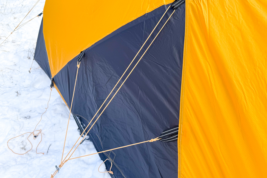 Close-up of the Mountain 25 fly showing taut guylines anchored in snow, with stitching and panel reinforcements visible on the yellow and black fabric.