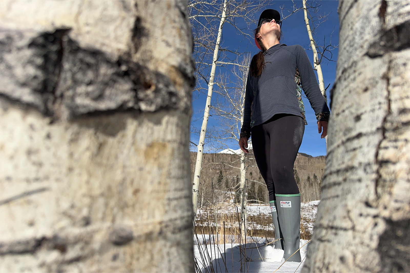 Two aspen trees frame a hiker wearing Xtratuf boots who is standing in the snow.