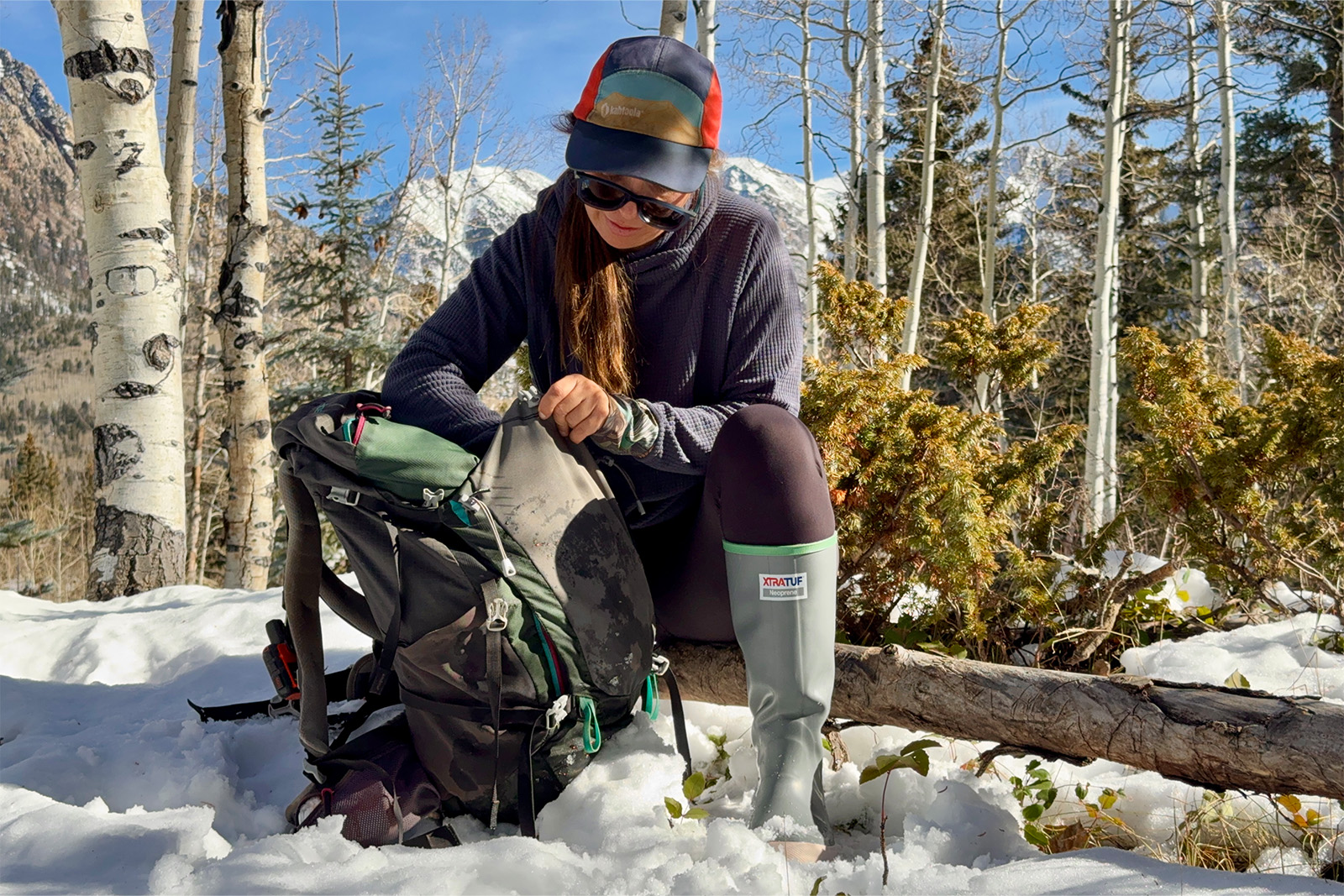 A person wearing Xtratuf boots is sitting on a log in the forest while pulling an item out of a backpack. There is snow on the ground.