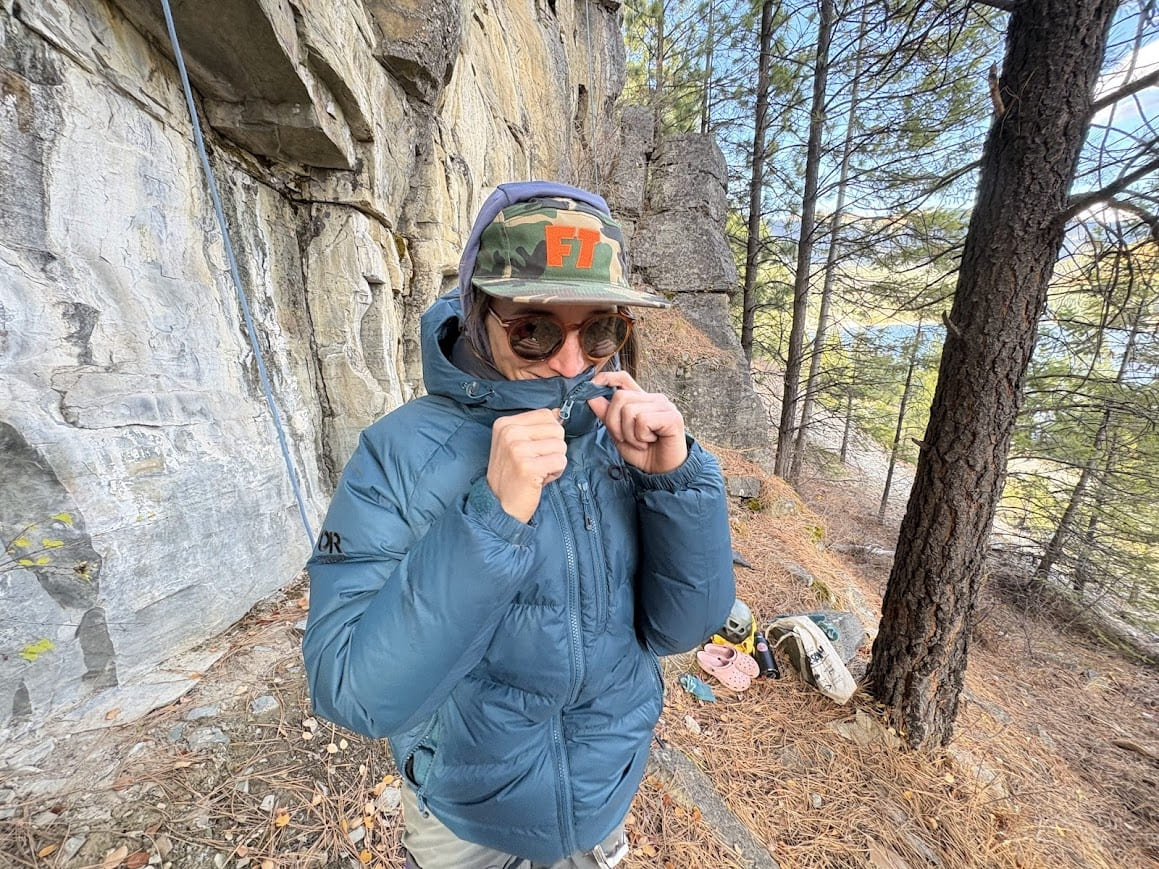 woman unzips a blue down jacket next to a rock climbing wall