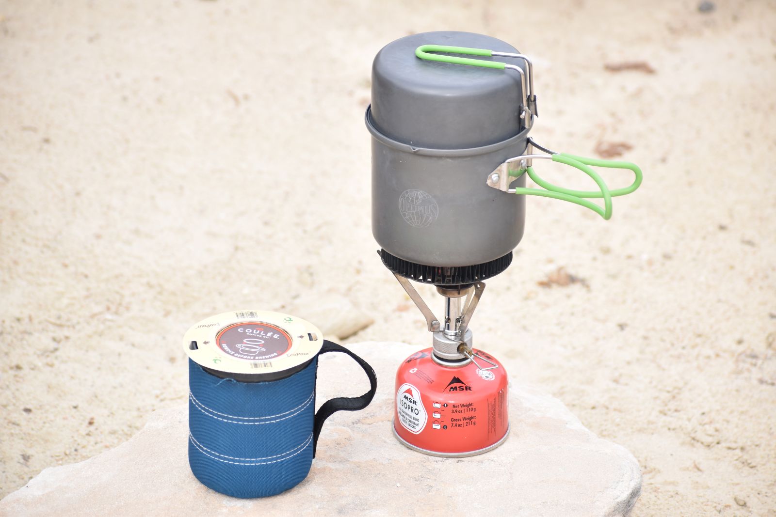 An EcoPour filter sits on a blue coffee mug next to an MSR Pocket Rocket camp stove