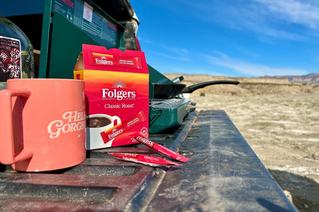 Up-close image of a box of Folgers instant coffee on a tailgate of a truck next to a mug and a camp stove
