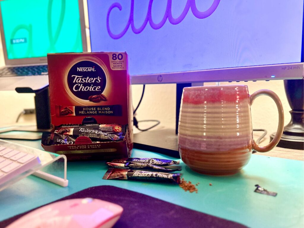 Up-close image of a large box of Nescafe Taster's Choice instant coffee packets with a mug next to it at a desk with computer monitors in the background