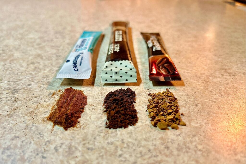 Up-close image of 3 different piles of instant coffee on a counter in front of their respective brand packaging.