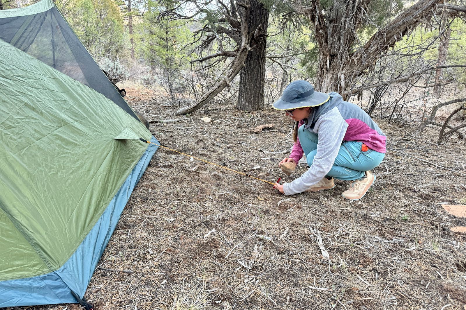 A person is using a rock to pound a tent stake into the ground for the side guy out line of the River Country trekker tent. There are pinion and juniper trees in the background.