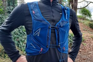 Close-up image of a woman wearing a blue running vest with a forest trail in the background.