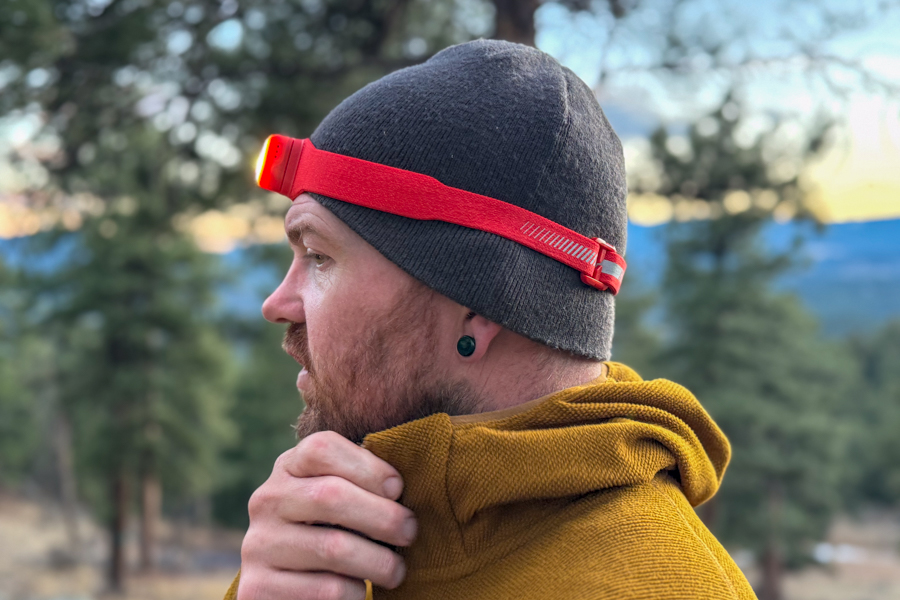 Side profile of a person wearing a red headlamp over a beanie at dusk in a pine forest.