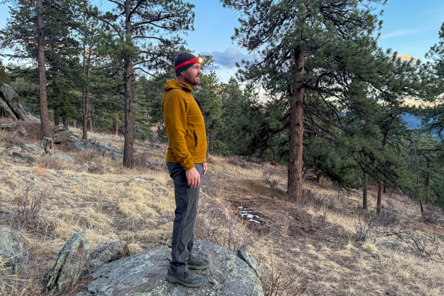 Wide shot of a person standing on a rocky hillside at dusk wearing a red headlamp in a pine forest.