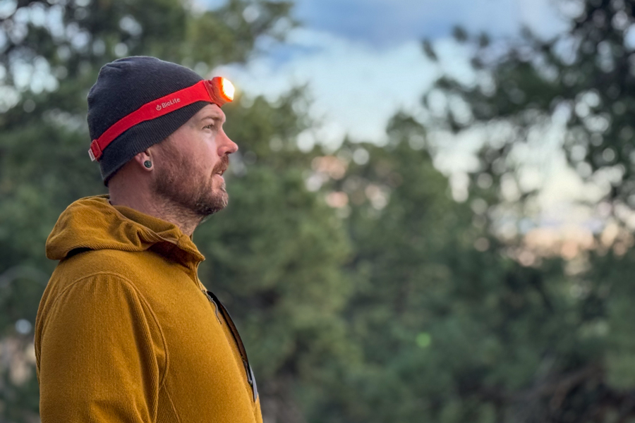 Side profile of a person wearing a red headlamp while standing outdoors at dusk with trees in the background.