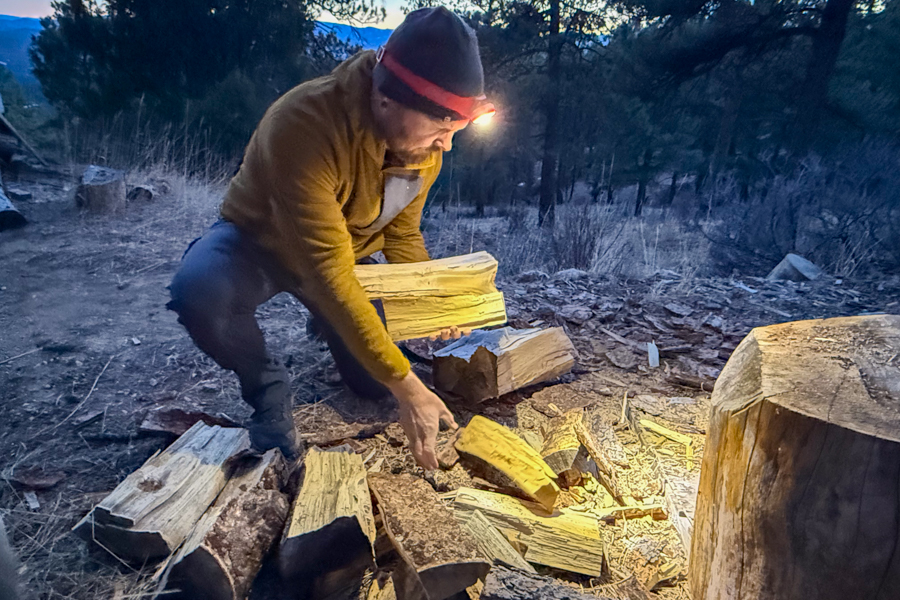 Person crouched near a woodpile at dusk, wearing a headlamp while arranging firewood.
