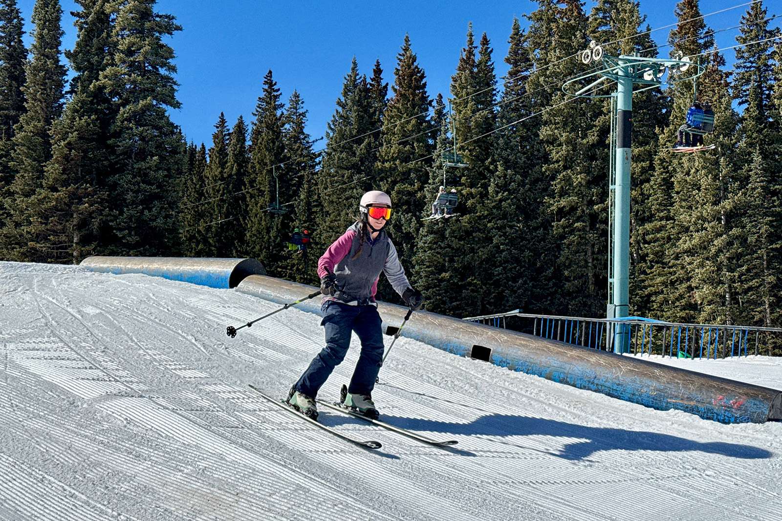 A skier using the cascade poles is skiing down a run next to some rails with a ski lift and some trees in the background.