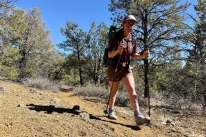 A person wearing a backpack and using the cascade poles is walking up a trail towards the camera. There are trees juniper trees in the background.