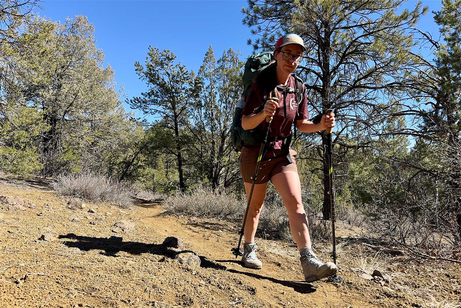 A person wearing a backpack and using the cascade poles is walking up a trail towards the camera. There are trees juniper trees in the background.
