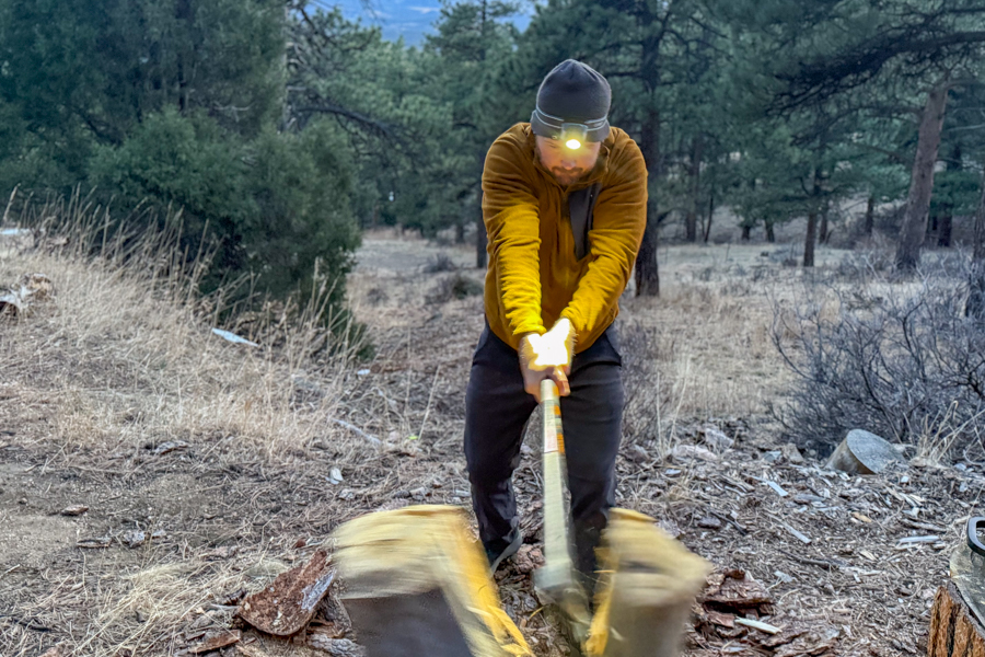 Person splitting wood with an axe at dusk while wearing a bright headlamp in a pine forest.