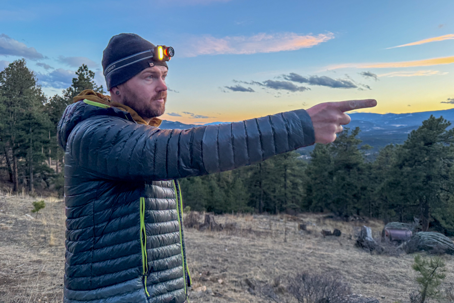 Person wearing a headlamp at sunset pointing into the distance on a hillside above a forested valley.