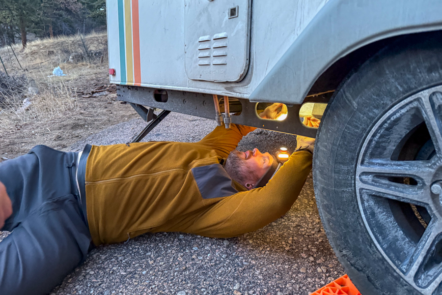 Person lying on the ground working underneath a trailer, using a headlamp to illuminate the underside near a wheel.
