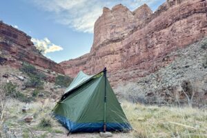 The Trekker tent is set up at the bottom of a canyon with high, red cliff walls surrounding it. The view is showing the back of the tent with a trekking pole easily visible holding it up.