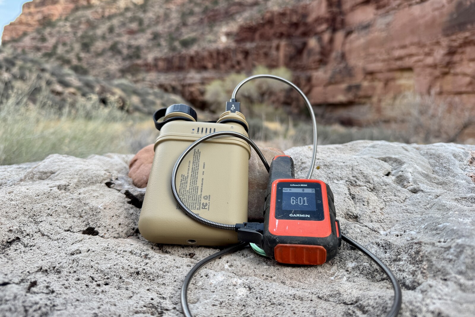 Close up of the Nestout charging a Garmin Inreach Mini while both are sitting on a rock. There is a red rock canyon in the background.