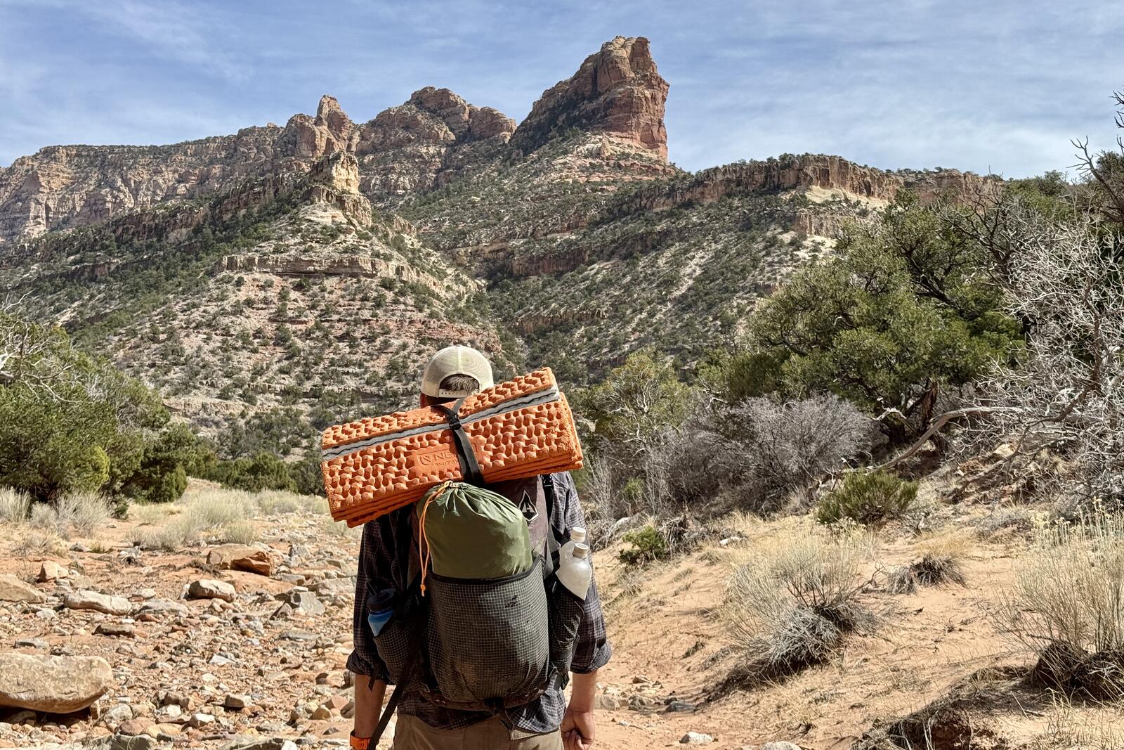 A person wearing a backpack is walking in a dry river wash with red rock formations in the distance. There is a foldable sleeping pad on top of the pack and a green bag stuffed in the back pocket which holds the River Country tent.