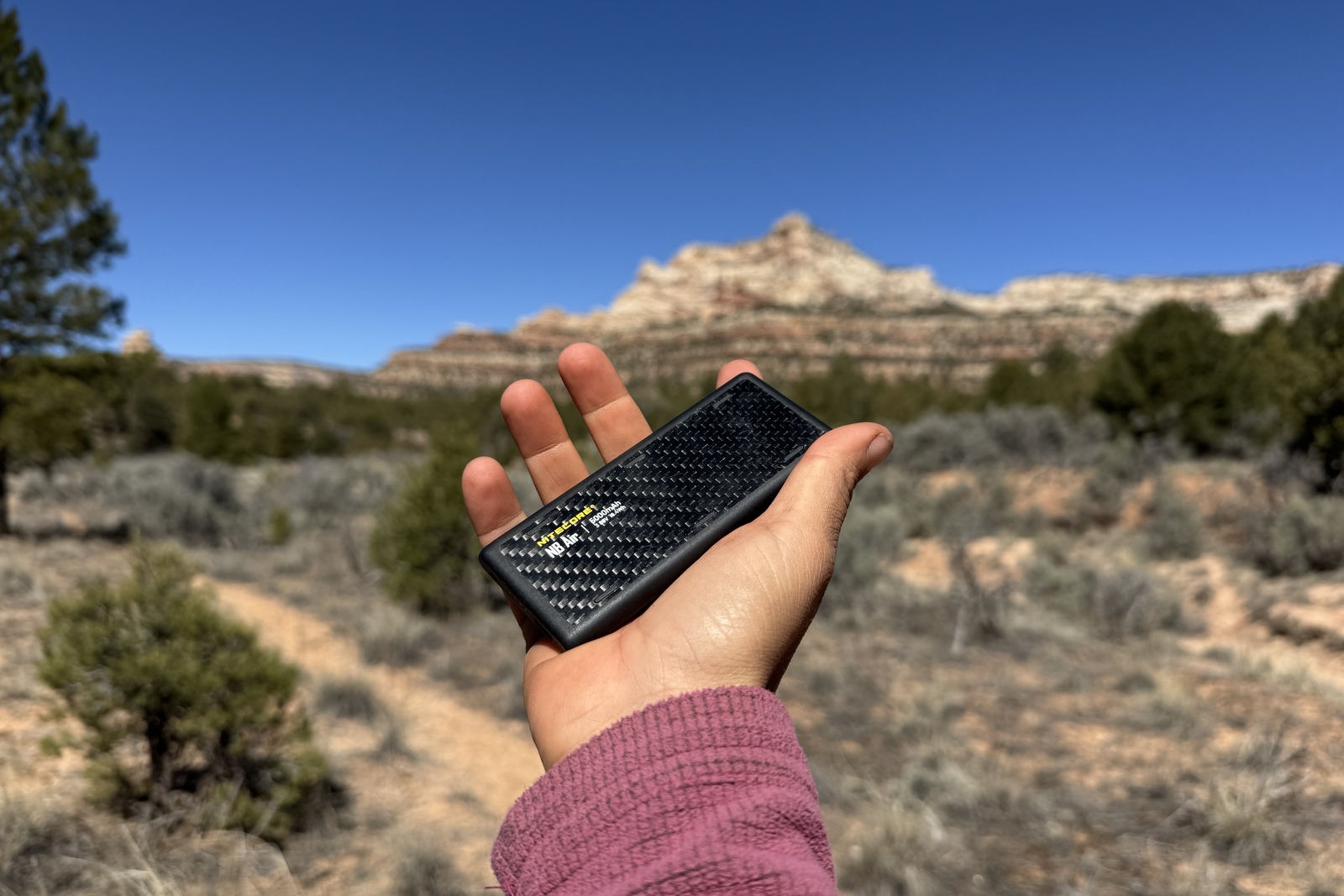Closeup of a hand holding out the NB Air. There are pinions and a red rock formation in the background.