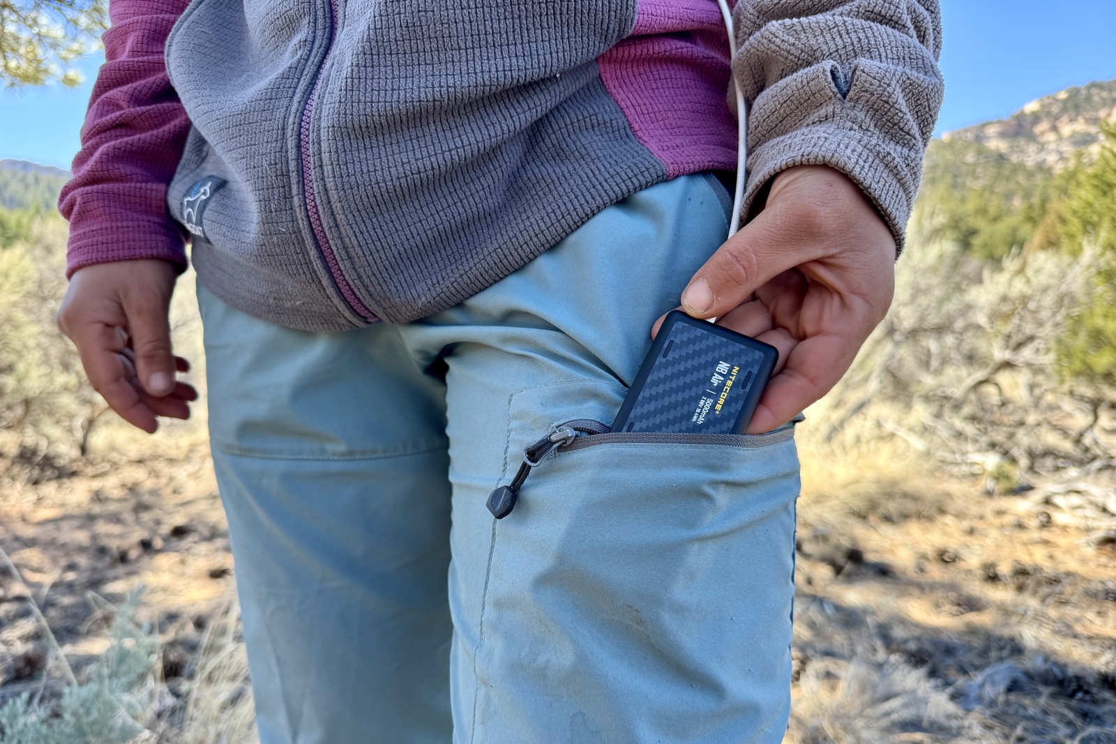 Closeup of a person putting the NB Air into a pants pocket. The NB Air is plugged into a charging cord and there is a pinion-juniper forest in the background.