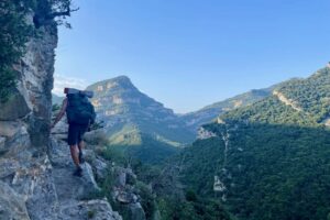 a hiker traverses a narrow ledge over a cliff with green mountains in the background