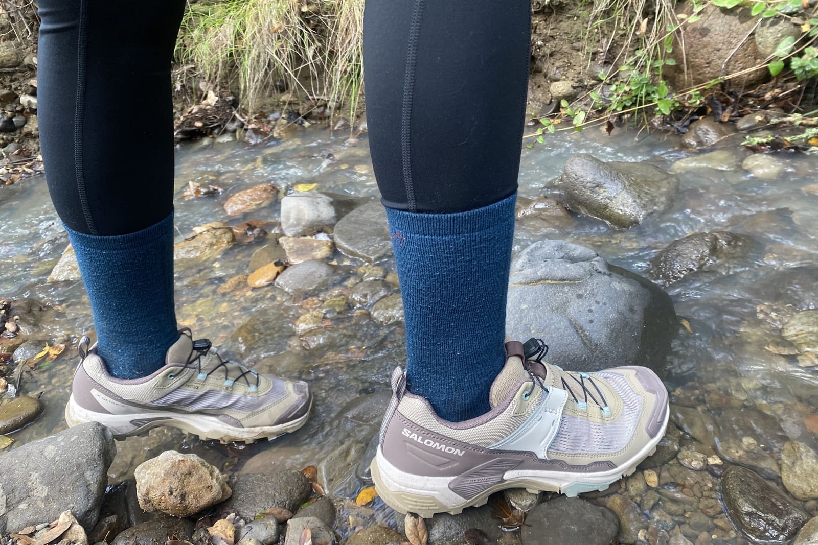 A woman stands along a creek in her shoes.