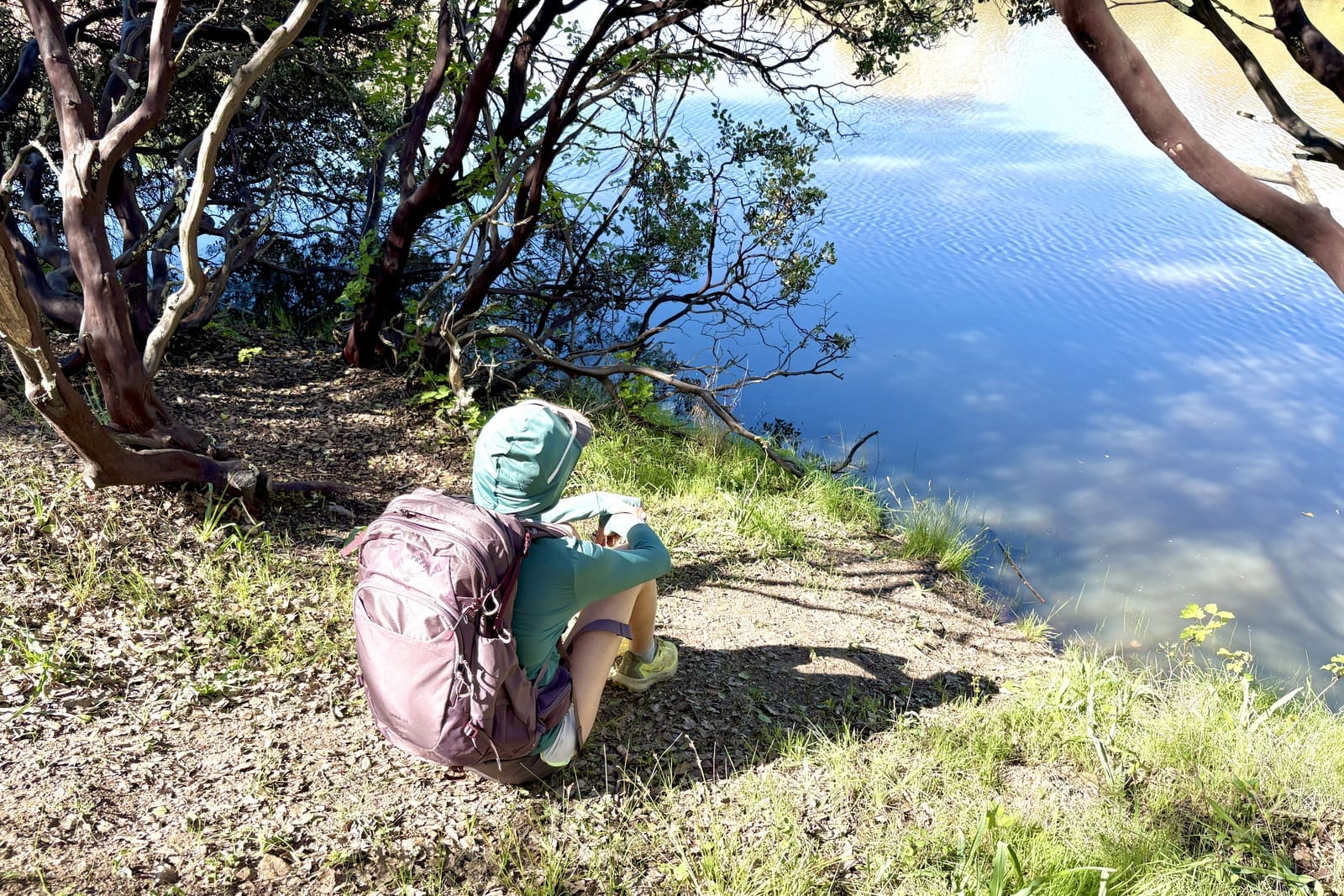 A woman sits next to a lake wearing her day pack.