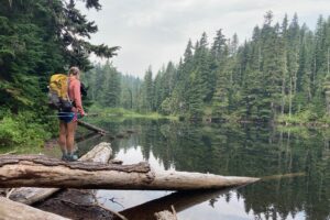 a hiker stands on a log next to a pond with a yellow backpack on her back
