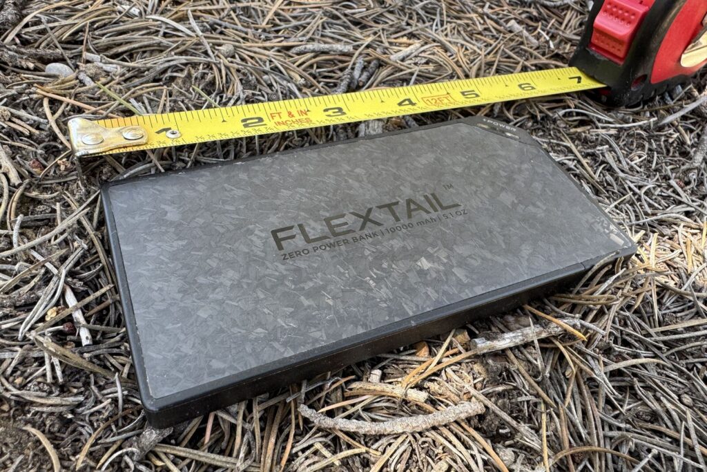 A close up of the Flextail power bank sitting on some pine needles with a measuring tape extended next to it.