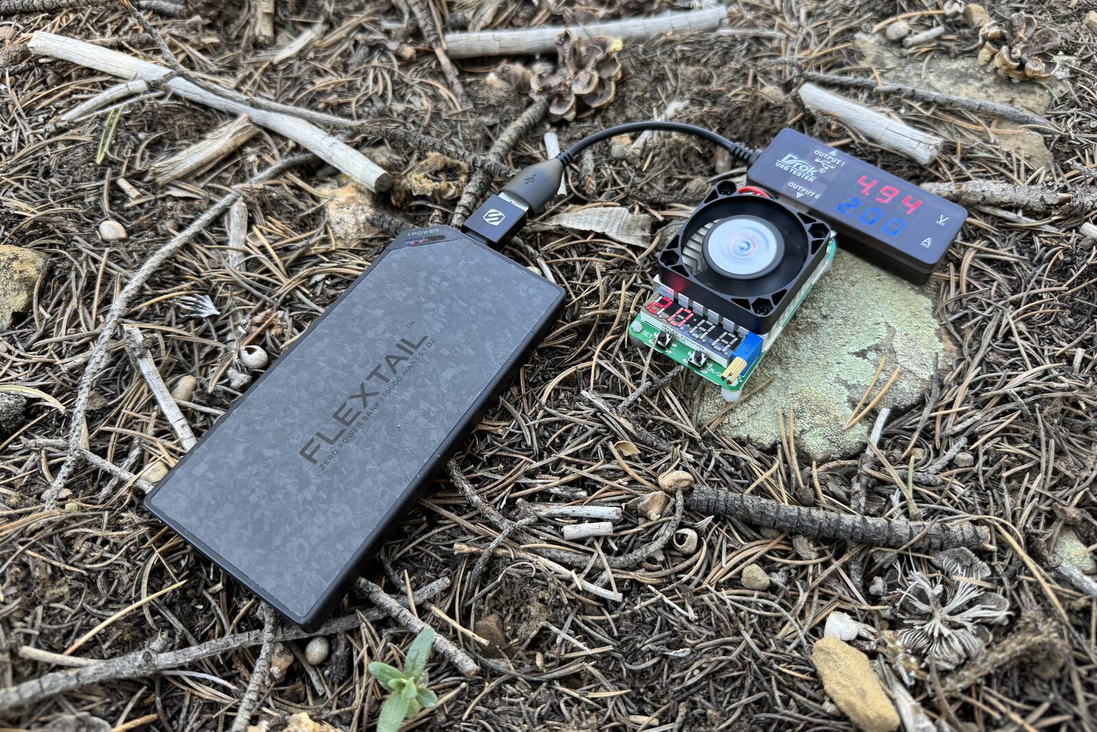 Close up of the Flextail power bank plugged into a multi-meter and dummy load resistor. All three are sitting on some pine needles on the ground.