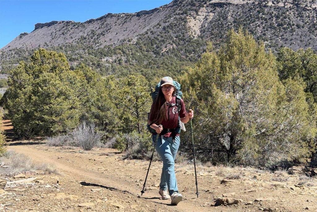 A person using the Legacy poles and wearing a pack is walking up a trail towards the camera with pinion and juniper trees and a mesa in the background.