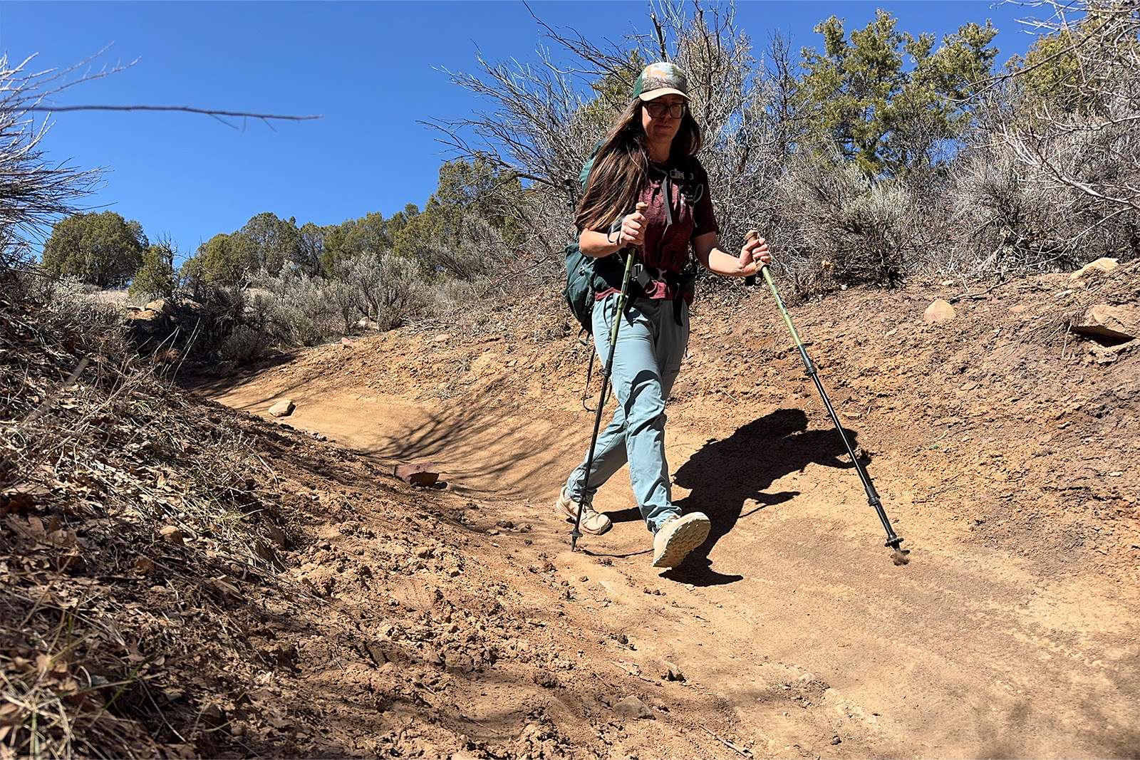 A person wearing a pack is using the Legacy poles to hike down a dirt trail with pinion and juniper trees in the background.