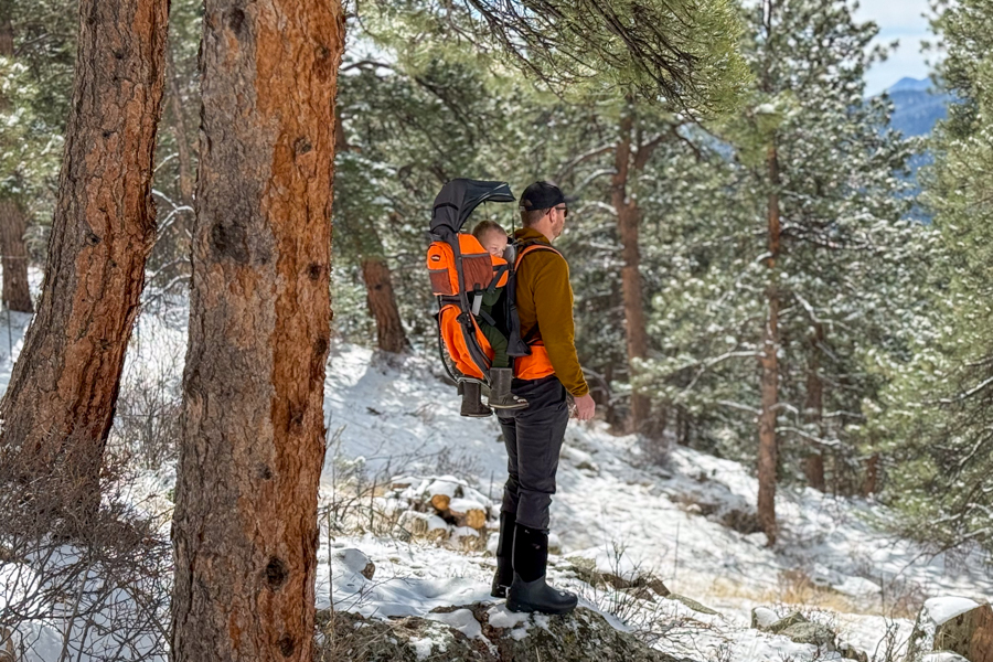 Tester standing on a snowy trail wearing the Luvdbaby Premium child carrier with a child riding in the seat beneath the deployed sunshade.