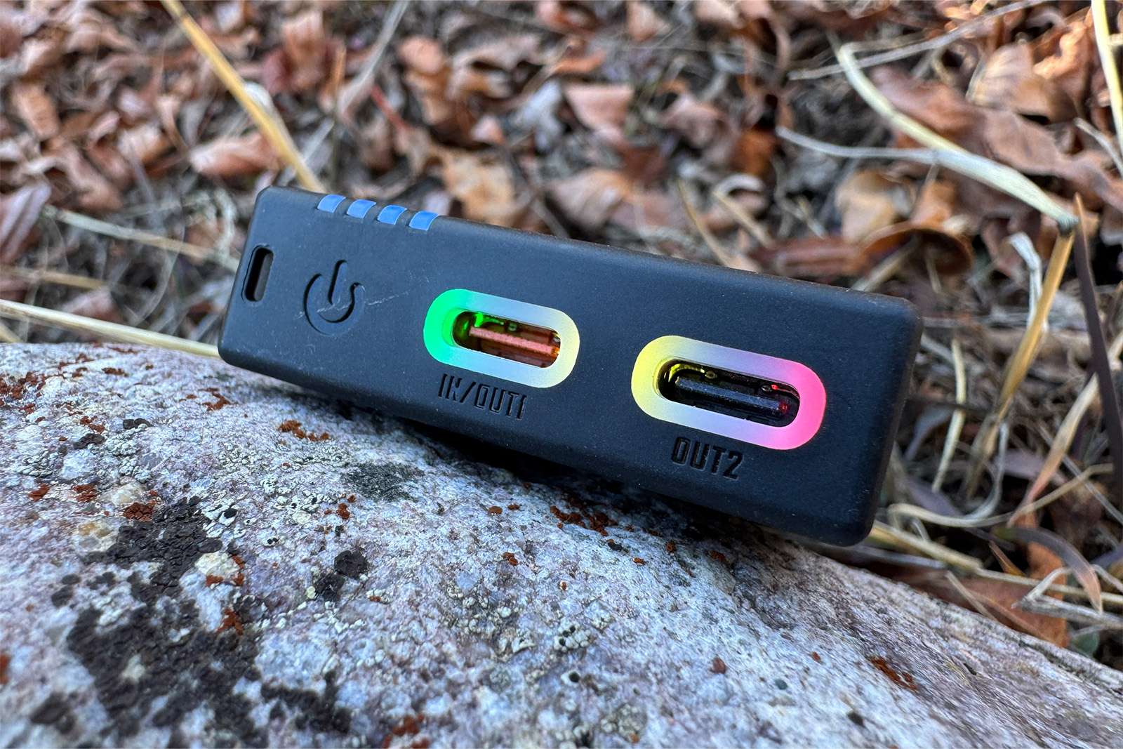 Close up of the NB10000 power bank propped up on a rock with leaves in the background. The ports are glowing a rainbow color with a power symbol next to them and a small charge indicator.
