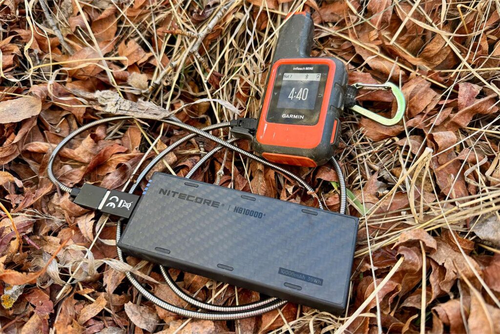 El NB10000 tiene un cable conectado y también conectado a un Garmin Inreach Mini. Ambos están sentados sobre unas hojas en el suelo.