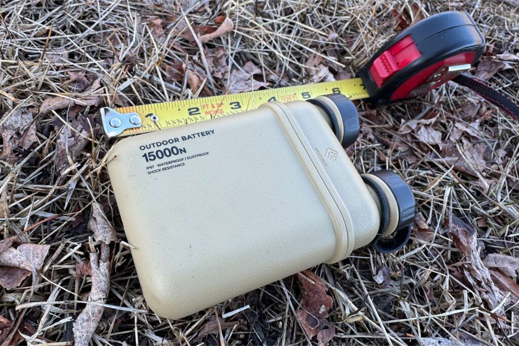 The Nestout power bank is laying on some leaves and grass on the ground with a measure tape extended next to it.