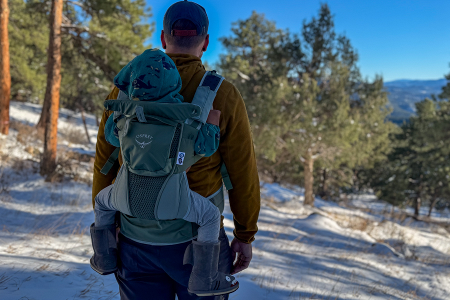 Tester hiking in snowy conditions with a child riding in the Osprey Poco Soft, showing the carrier’s back panel and compact shape.