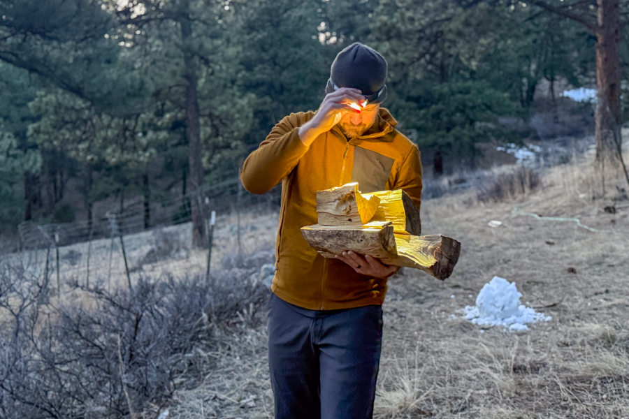 Person carrying a stack of firewood at dusk while using a headlamp with a red light mode.
